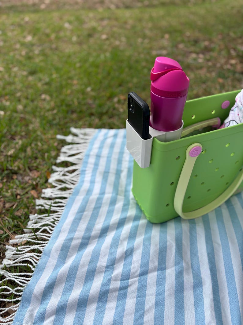 Op de afbeelding: Een limoengroene draagtas met een witte telefoonhouder, een magenta waterfles en een zwarte smartphone. De tas staat op een blauw-wit gestreepte deken met een gefranjerde rand, op een grasachtig oppervlak.