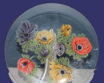 May include: A clear glass decorative plate featuring a floral design with anemone flowers in shades of red, yellow, purple, and white. The flowers are surrounded by green leaves. The plate is displayed on a silver stand.