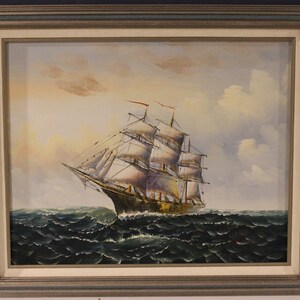 May include: Oil painting of a tall ship sailing on a wavy sea under a cloudy sky. The ship has multiple white sails and red flags. The painting is in a silver frame.