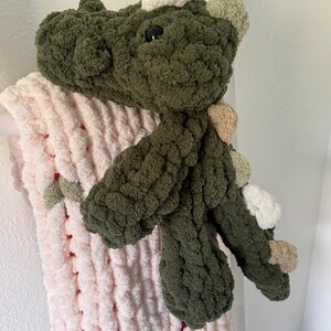 K&ouml;nnte beinhalten: Ein handgefertigtes, gr&uuml;nes geh&auml;keltes Dinosaurierspielzeug mit schwarzen Augen und verschiedenen farbigen Akzenten. Der Dinosaurier liegt auf einer weichen, rosa, grob gestrickten Decke. Das Spielzeug hat ein strukturiertes, pl&uuml;schiges Aussehen.