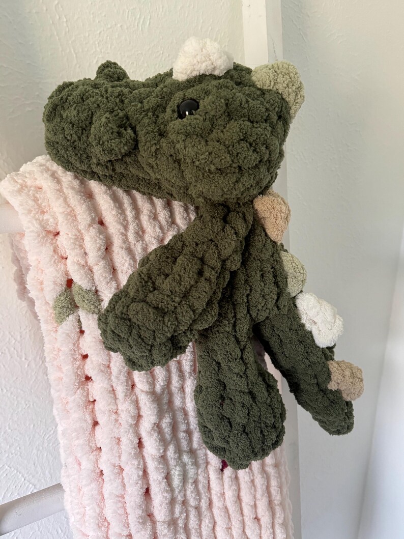 K&ouml;nnte beinhalten: Ein handgefertigtes, gr&uuml;nes geh&auml;keltes Dinosaurierspielzeug mit schwarzen Augen und verschiedenen farbigen Akzenten. Der Dinosaurier liegt auf einer weichen, rosa, grob gestrickten Decke. Das Spielzeug hat ein strukturiertes, pl&uuml;schiges Aussehen.