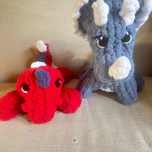 K&ouml;nnte beinhalten: Zwei handgefertigte Dinosaurier-Spielzeuge aus H&auml;kelarbeit. Einer ist ein roter Dinosaurier mit wei&szlig;en und lila Akzenten, der andere ein blauer Triceratops mit wei&szlig;en H&ouml;rnern und Akzenten. Beide haben schwarze Augen.