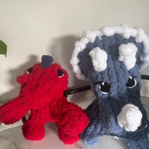 K&ouml;nnte beinhalten: Zwei handgefertigte Dinosaurier-Spielzeuge aus H&auml;kelarbeit. Eines ist rot mit einem dunkelblauen Akzent, das andere blau mit wei&szlig;en Akzenten. Beide haben schwarze Augen und sitzen auf einer wei&szlig;en Oberfl&auml;che.