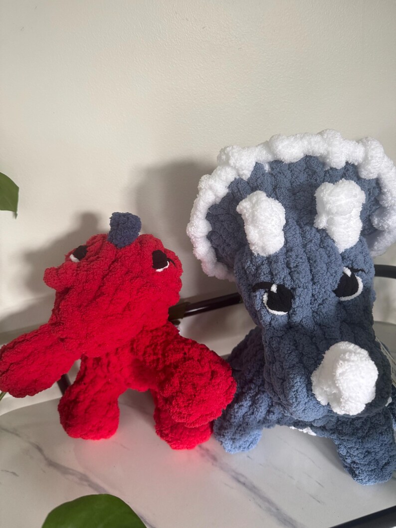 K&ouml;nnte beinhalten: Zwei handgefertigte Dinosaurier-Spielzeuge aus H&auml;kelarbeit. Eines ist rot mit einem dunkelblauen Akzent, das andere blau mit wei&szlig;en Akzenten. Beide haben schwarze Augen und sitzen auf einer wei&szlig;en Oberfl&auml;che.