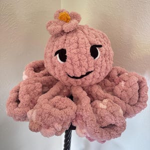 May include: A handmade pink plush octopus with a smiling face, black eyes, and a flower on its head. The octopus has eight curled tentacles and is made of a soft, textured material. The flower has a yellow center.