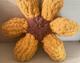 May include: A plush flower-shaped pillow crafted from chunky, textured yarn. The center is a dark brown circular shape, surrounded by six rounded, golden-yellow petals. The soft, knitted material has a thick, looped appearance throughout.
