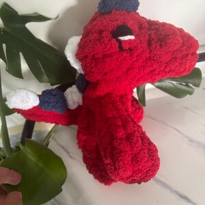 K&ouml;nnte beinhalten: Ein handgefertigtes, rotes geh&auml;keltes Dinosaurier-Pl&uuml;schtier mit einem blau-wei&szlig;en Schal und einem blauen Hut. Der Dinosaurier hat schwarze Augen und wei&szlig;e Akzente. Das Pl&uuml;schtier ist aus einem weichen, strukturierten Garn gefertigt.