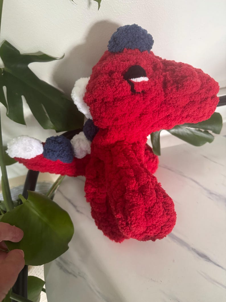 K&ouml;nnte beinhalten: Ein handgefertigtes, rotes geh&auml;keltes Dinosaurier-Pl&uuml;schtier mit einem blau-wei&szlig;en Schal und einem blauen Hut. Der Dinosaurier hat schwarze Augen und wei&szlig;e Akzente. Das Pl&uuml;schtier ist aus einem weichen, strukturierten Garn gefertigt.