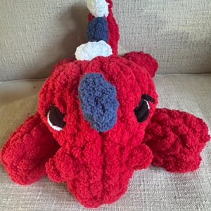 K&ouml;nnte beinhalten: Ein handgefertigter roter H&auml;kchendrache mit blauen und wei&szlig;en Akzenten. Der Drache hat schwarze Augen und liegt. Der Schwanz hat wei&szlig;e und blaue Pompons.