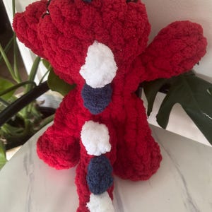 K&ouml;nnte beinhalten: Ein handgefertigtes, rotes geh&auml;keltes Drachen-Pl&uuml;schtier mit wei&szlig;en und marineblauen Pompon-Akzenten. Der Drache hat kleine schwarze Augen und ein strukturiertes, flauschiges Aussehen. Das Spielzeug ist als weiches, kuscheliges Objekt konzipiert.