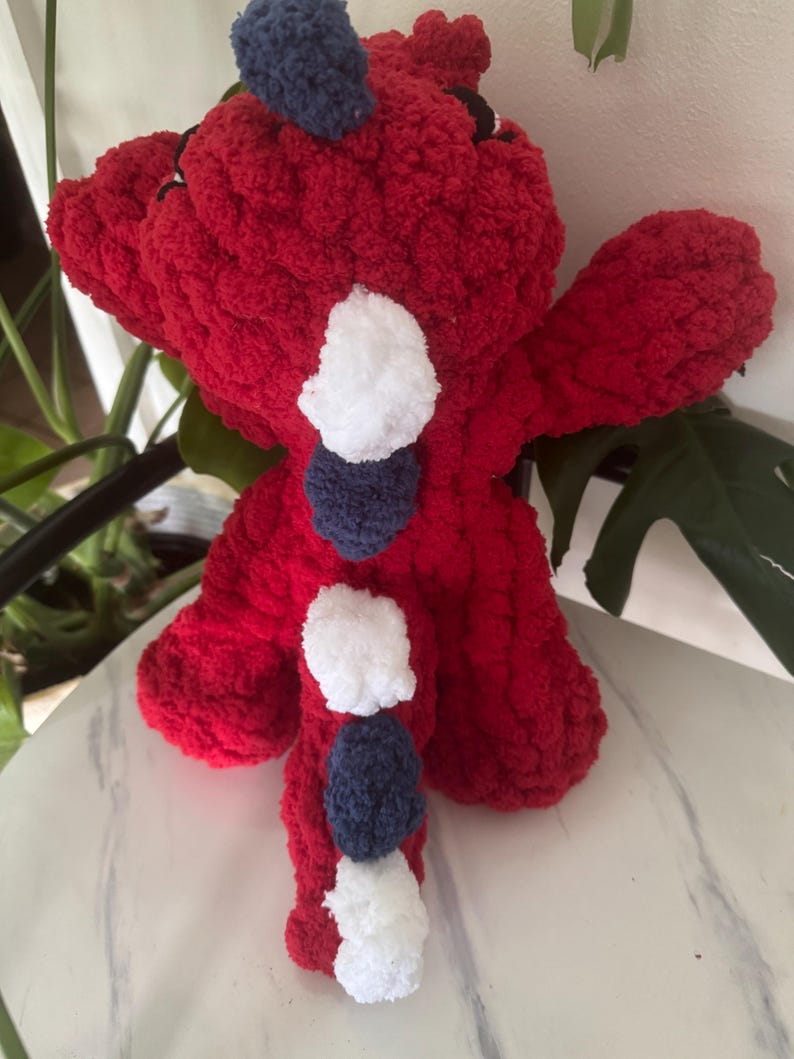 K&ouml;nnte beinhalten: Ein handgefertigtes, rotes geh&auml;keltes Drachen-Pl&uuml;schtier mit wei&szlig;en und marineblauen Pompon-Akzenten. Der Drache hat kleine schwarze Augen und ein strukturiertes, flauschiges Aussehen. Das Spielzeug ist als weiches, kuscheliges Objekt konzipiert.