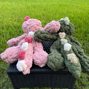 K&ouml;nnte beinhalten: Zwei handgefertigte, pl&uuml;schige Oktopus-Spielzeuge. Eines ist rosa mit roten und wei&szlig;en Pompons, das andere ist gr&uuml;n mit beige-wei&szlig;en Akzenten. Die Spielzeuge sind aus einem groben, weichen Garn gefertigt und liegen auf einem schwarzen Korbstuhl.