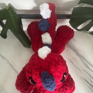 K&ouml;nnte beinhalten: Ein handgefertigtes rotes Pl&uuml;schtier mit wei&szlig;en und marineblauen Akzenten. Das Spielzeug hat ein strukturiertes, flauschiges Aussehen mit kleinen schwarzen Augen und einer marineblauen Nase. Das Design des Spielzeugs umfasst wei&szlig;e und marineblaue Pompons auf dem R&uuml;cken.