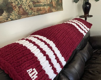 May include: A chunky knit throw blanket in deep maroon with two thick white horizontal stripes. The blanket features the white embroidered letters ATM in a collegiate font on the lower corner, showcasing a soft, textured yarn construction.