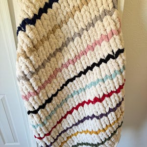 May include: A chunky knit blanket in cream with diagonal stripes of various colors, including navy, pink, black, and red. The blanket is draped over a white ladder, showcasing its texture and design. This cozy blanket is perfect for home decor.