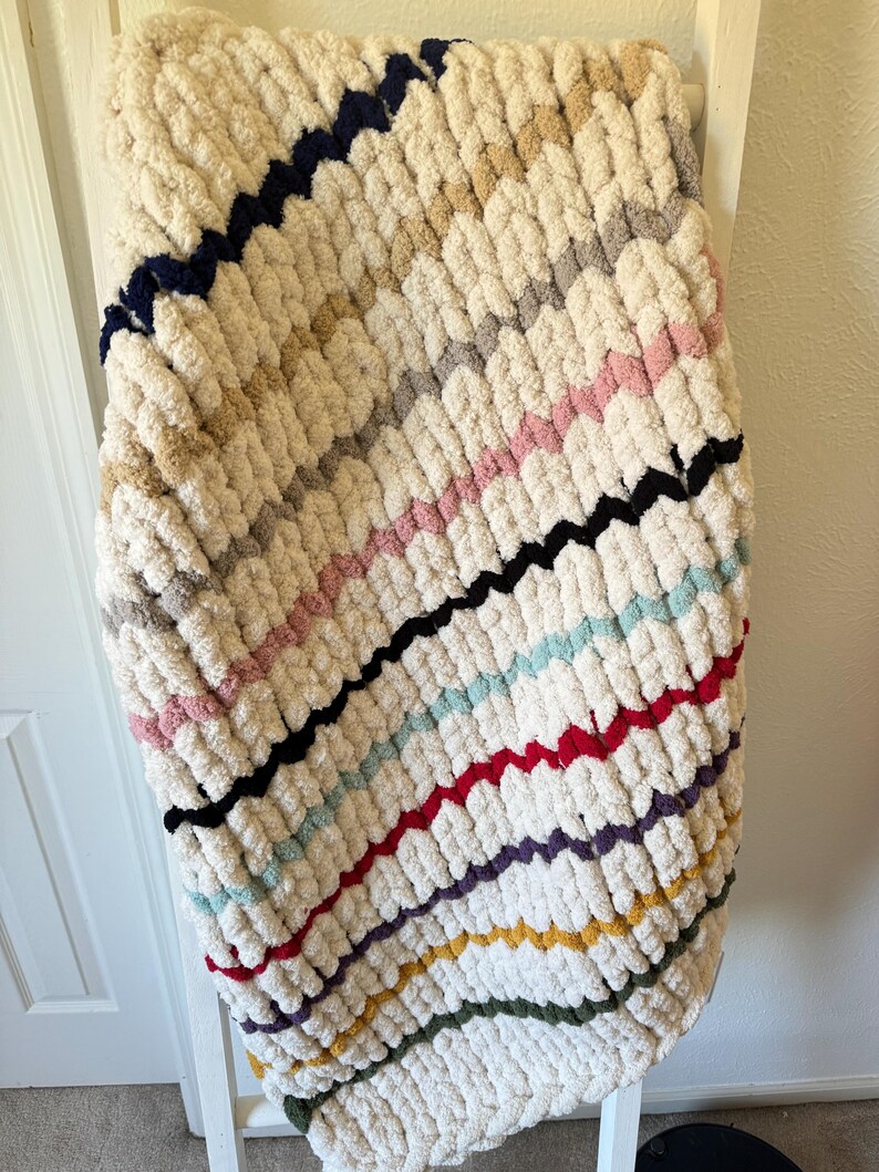 May include: A chunky knit blanket in cream with diagonal stripes of various colors, including navy, pink, black, and red. The blanket is draped over a white ladder, showcasing its texture and design. This cozy blanket is perfect for home decor.