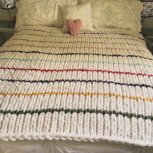 May include: A cream-colored, chunky knit blanket with horizontal stripes in various colors, including red, blue, yellow, and green. A pink heart-shaped pillow sits on top of the blanket, which is on a bed with decorative pillows.