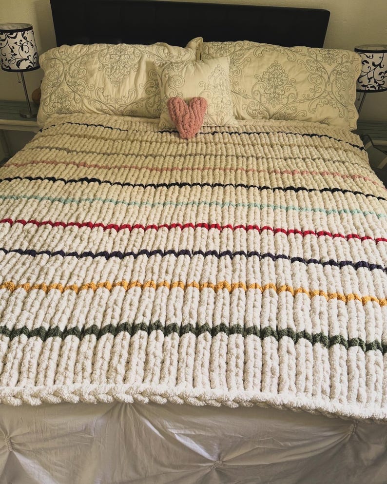 May include: A cream-colored, chunky knit blanket with horizontal stripes in various colors, including red, blue, yellow, and green. A pink heart-shaped pillow sits on top of the blanket, which is on a bed with decorative pillows.