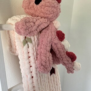 K&ouml;nnte beinhalten: Ein handgefertigtes, pl&uuml;schiges rosa Drachenspielzeug mit passender Decke. Der Drache hat einen hellrosa K&ouml;rper, dunklere rosa Akzente und schwarze Augen. Die Decke ist cremefarben mit rosa Akzenten und Blattdetails.