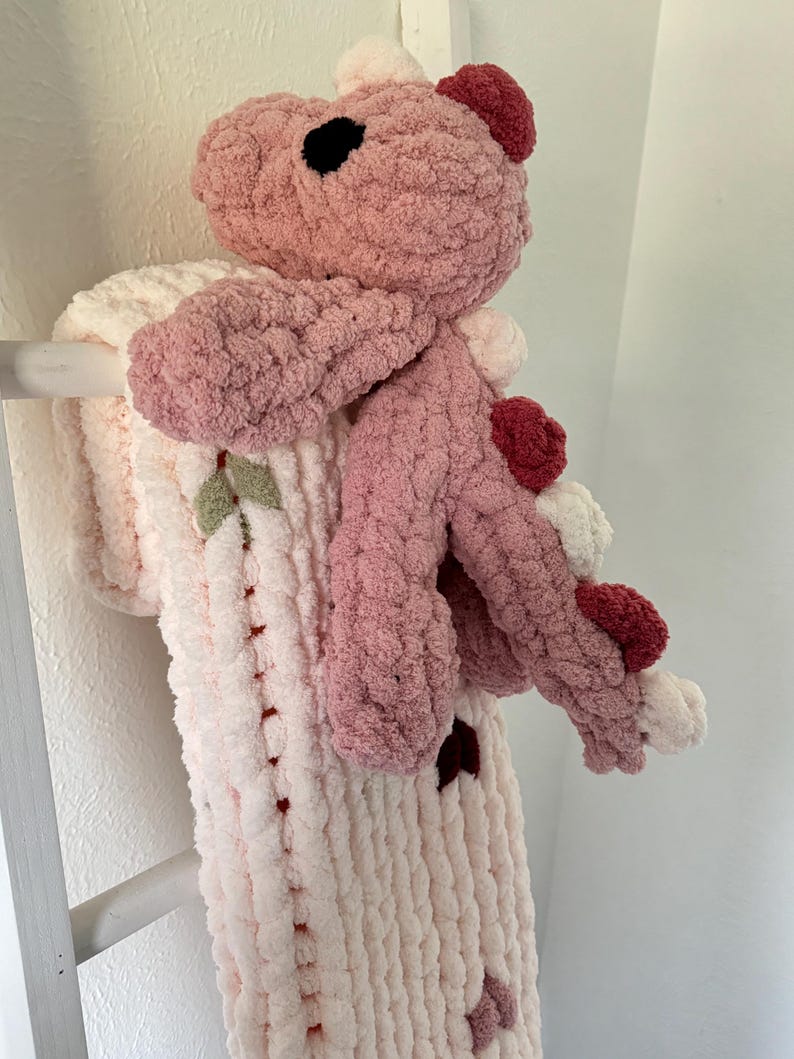 K&ouml;nnte beinhalten: Ein handgefertigtes, pl&uuml;schiges rosa Drachenspielzeug mit passender Decke. Der Drache hat einen hellrosa K&ouml;rper, dunklere rosa Akzente und schwarze Augen. Die Decke ist cremefarben mit rosa Akzenten und Blattdetails.