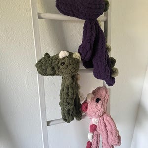 K&ouml;nnte beinhalten: Drei handgefertigte Dinosaurier-Pl&uuml;schtiere in Lila, Gr&uuml;n und Rosa, jeweils mit kontrastierenden Akzenten. Die Pl&uuml;schtiere werden auf einer wei&szlig;en Leiter pr&auml;sentiert, die ihr weiches, strukturiertes Aussehen zeigt. Die Dinosaurier haben kleine, runde Augen und dekorative Stacheln.