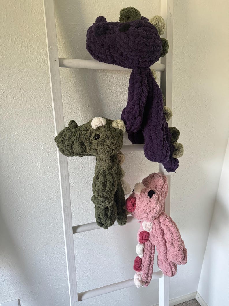 K&ouml;nnte beinhalten: Drei handgefertigte Dinosaurier-Pl&uuml;schtiere in Lila, Gr&uuml;n und Rosa, jeweils mit kontrastierenden Akzenten. Die Pl&uuml;schtiere werden auf einer wei&szlig;en Leiter pr&auml;sentiert, die ihr weiches, strukturiertes Aussehen zeigt. Die Dinosaurier haben kleine, runde Augen und dekorative Stacheln.