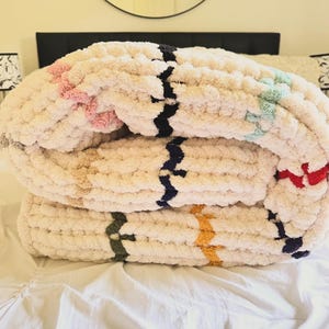 May include: A cream-colored, chunky knit blanket folded on a bed. The blanket features stripes of pink, black, blue, green, yellow, and red. The blanket is made of a soft, thick yarn, perfect for adding warmth and texture to a bedroom.