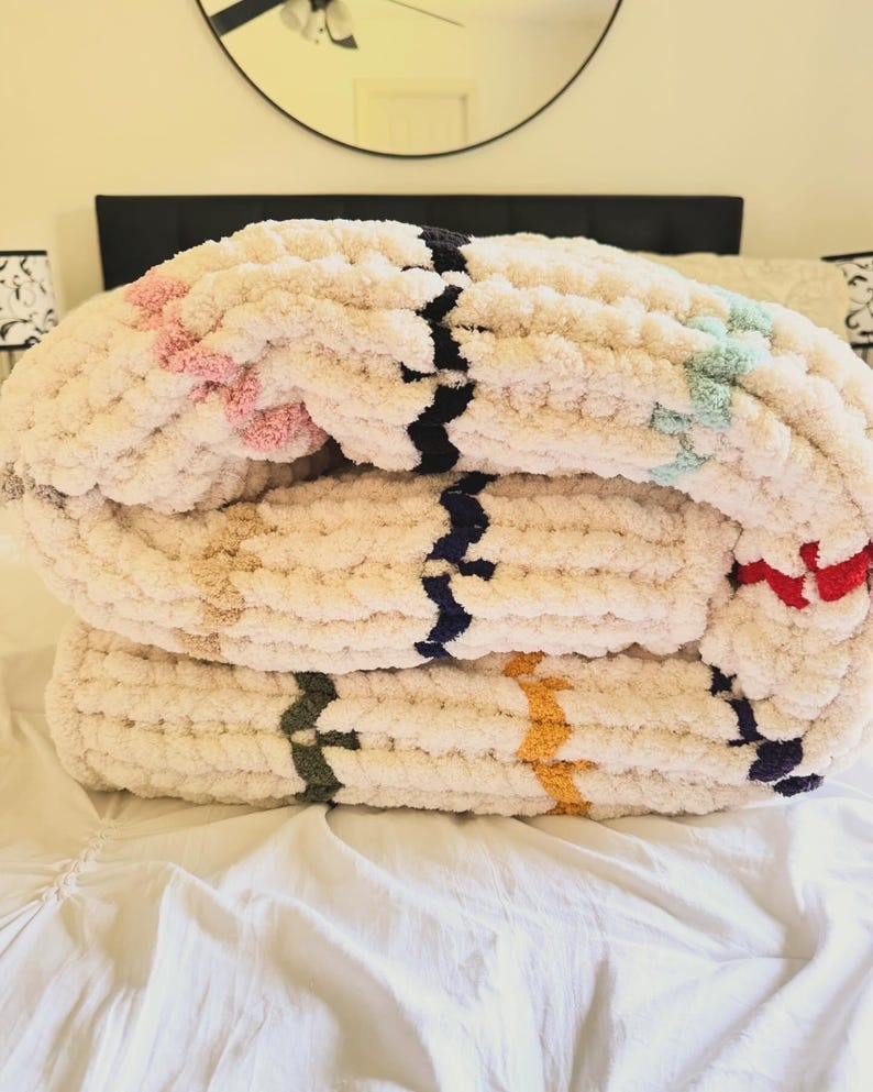 May include: A cream-colored, chunky knit blanket folded on a bed. The blanket features stripes of pink, black, blue, green, yellow, and red. The blanket is made of a soft, thick yarn, perfect for adding warmth and texture to a bedroom.