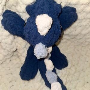 K&ouml;nnte beinhalten: Ein handgefertigtes, pl&uuml;schiges Dinosaurierspielzeug in Marineblau und Wei&szlig;. Der Dinosaurier hat ein flauschiges, strukturiertes Aussehen mit wei&szlig;en Akzenten auf Kopf und R&uuml;cken. Das Spielzeug befindet sich vor einem weichen, wei&szlig;en, strukturierten Hintergrund.