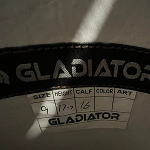 May include: A black interior label for a Gladiator brand product featuring a white tag with printed headers for SIZE, HEIGHT, CALF, COLOR, and ART. Handwritten entries include 9 for size, 17.7 for height, and 16 for calf, with the brand name Gladiator.