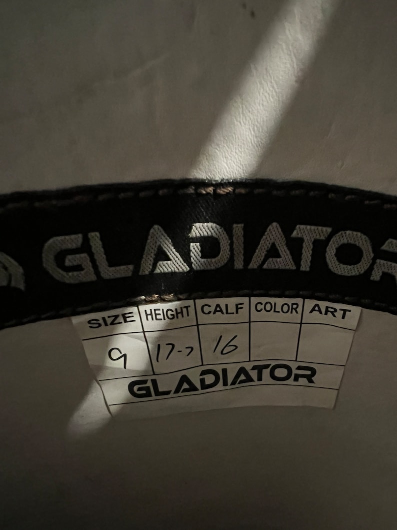 May include: A black interior label for a Gladiator brand product featuring a white tag with printed headers for SIZE, HEIGHT, CALF, COLOR, and ART. Handwritten entries include 9 for size, 17.7 for height, and 16 for calf, with the brand name Gladiator.
