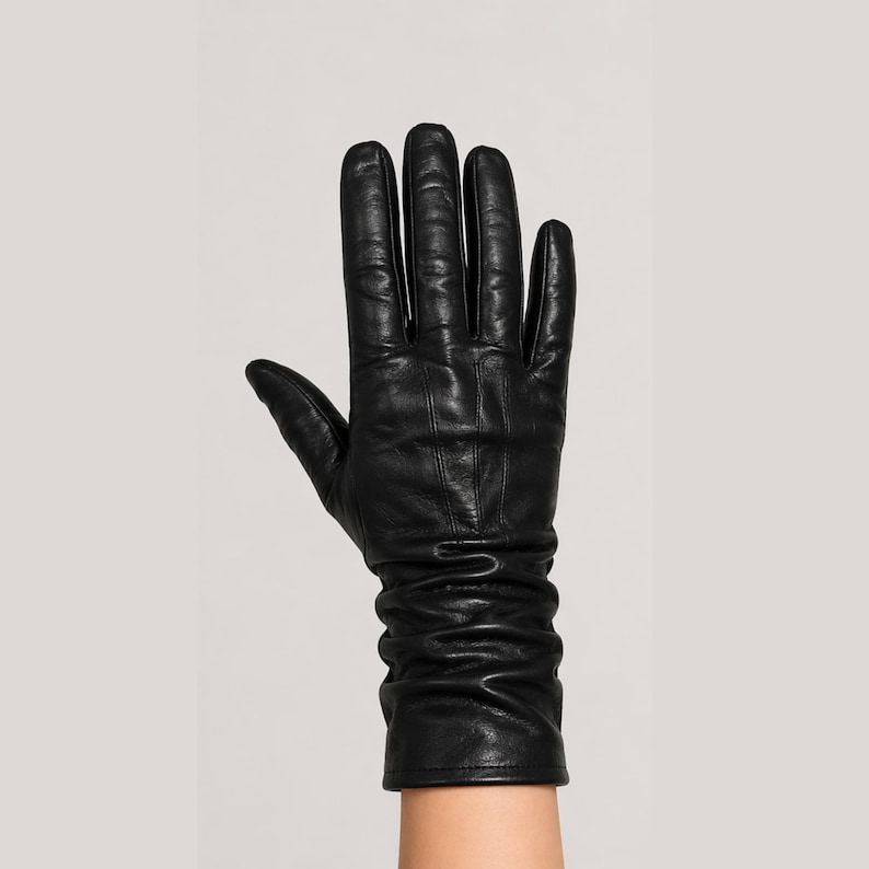 May include: A black leather glove with a smooth finish and visible stitching along the fingers. The wrist area features a gathered, ruched design that creates soft folds in the material, extending upward to cover the base of the wrist area.