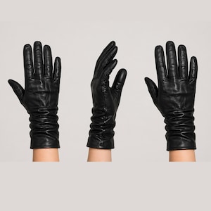 May include: A set of three black leather gloves shown from different angles. Each glove features a smooth finish, five fingers, and a gathered, ruched design at the wrist area. The leather has a slight sheen and visible natural texture throughout.