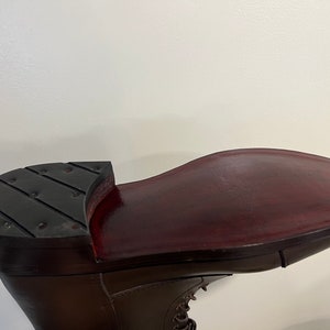 May include: A dark brown leather boot featuring a smooth, polished deep burgundy sole and a black heel cap with a series of small, evenly spaced metal studs embedded across the surface. The boot has a lace-up front and a structured silhouette.