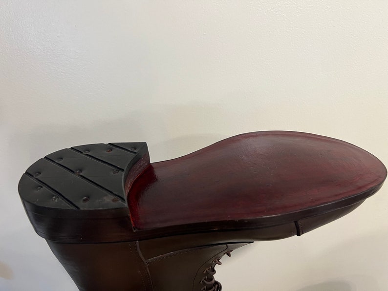 May include: A dark brown leather boot featuring a smooth, polished deep burgundy sole and a black heel cap with a series of small, evenly spaced metal studs embedded across the surface. The boot has a lace-up front and a structured silhouette.