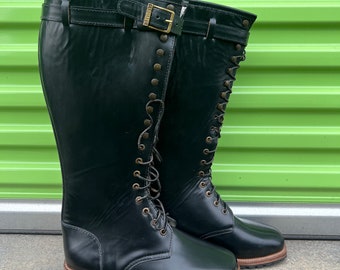 May include: A pair of black leather knee-high boots with laces and a buckle. The boots have a brown sole and are sitting on a concrete surface.