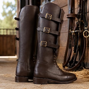 May include: A pair of dark brown leather tall riding boots featuring a lace-up front design, a rounded toe, and three horizontal straps across the upper shaft. Each strap is secured with a gold-toned metal buckle, and the boots have a low heel.