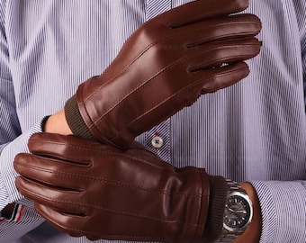May include: A pair of brown leather gloves featuring three decorative stitched lines on the back of the hand and a dark brown ribbed knit cuff at the wrist. The gloves have a smooth finish and are designed to cover the fingers and the wrist area.