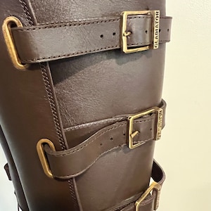 May include: A dark brown leather boot featuring three horizontal straps with gold-toned metal buckles. Each buckle is engraved with the word GLADIATOR. The straps are secured through vertical metal loops attached to the side of the boot shaft.