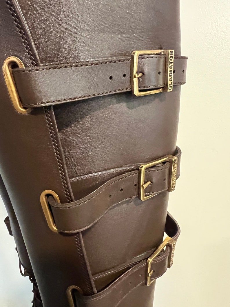 May include: A dark brown leather boot featuring three horizontal straps with gold-toned metal buckles. Each buckle is engraved with the word GLADIATOR. The straps are secured through vertical metal loops attached to the side of the boot shaft.