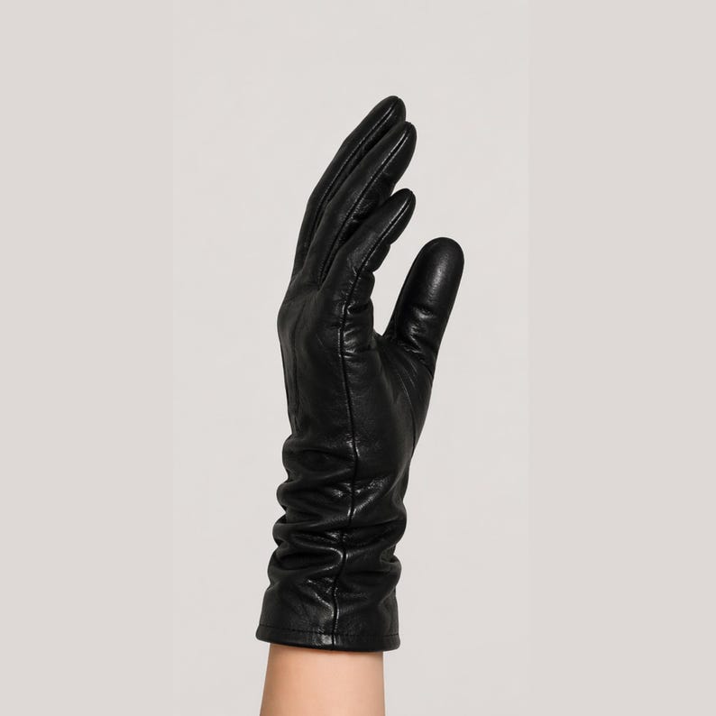 May include: A black leather glove with a smooth finish and a slightly ruched, gathered wrist area. The glove features visible stitching along the fingers and palm, extending to cover the wrist with a clean, straight-cut hem at the bottom edge.