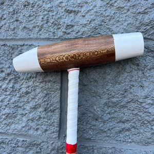 Könnte beinhalten: Ein Holz-Polo-Schläger mit einem weißen und braunen Kopf und einem weißen Griff. Der Schaft ist mit weißem Klebeband umwickelt und hat ein rotes Band.