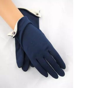 May include: A pair of navy blue gloves featuring a contrasting cream-colored cuff edge and a small, dark circular button detail at the wrist. The gloves are constructed from a smooth, solid-colored fabric and extend to cover the fingers and palm.