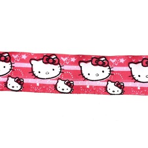 May include: Pink ribbon with a repeating pattern of white Hello Kitty faces with red bows. The ribbon has a white background with pink stripes and a white polka dot pattern.