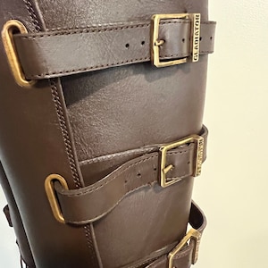 May include: A dark brown boot featuring three horizontal straps with brass-toned metal buckles. Each buckle is engraved with the word GLADIATOR. The boot has matching brass-toned metal loops securing the straps against the smooth brown surface.