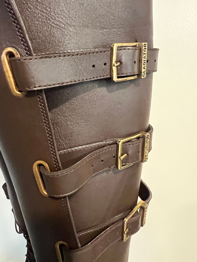 May include: A dark brown boot featuring three horizontal straps with brass-toned metal buckles. Each buckle is engraved with the word GLADIATOR. The boot has matching brass-toned metal loops securing the straps against the smooth brown surface.