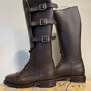 May include: A pair of dark brown knee-high boots featuring a lace-up front and a side panel secured by three horizontal straps with brass-toned buckles. Each buckle is embossed with the word GLADIATOR. The boots have a rounded toe and a low heel.