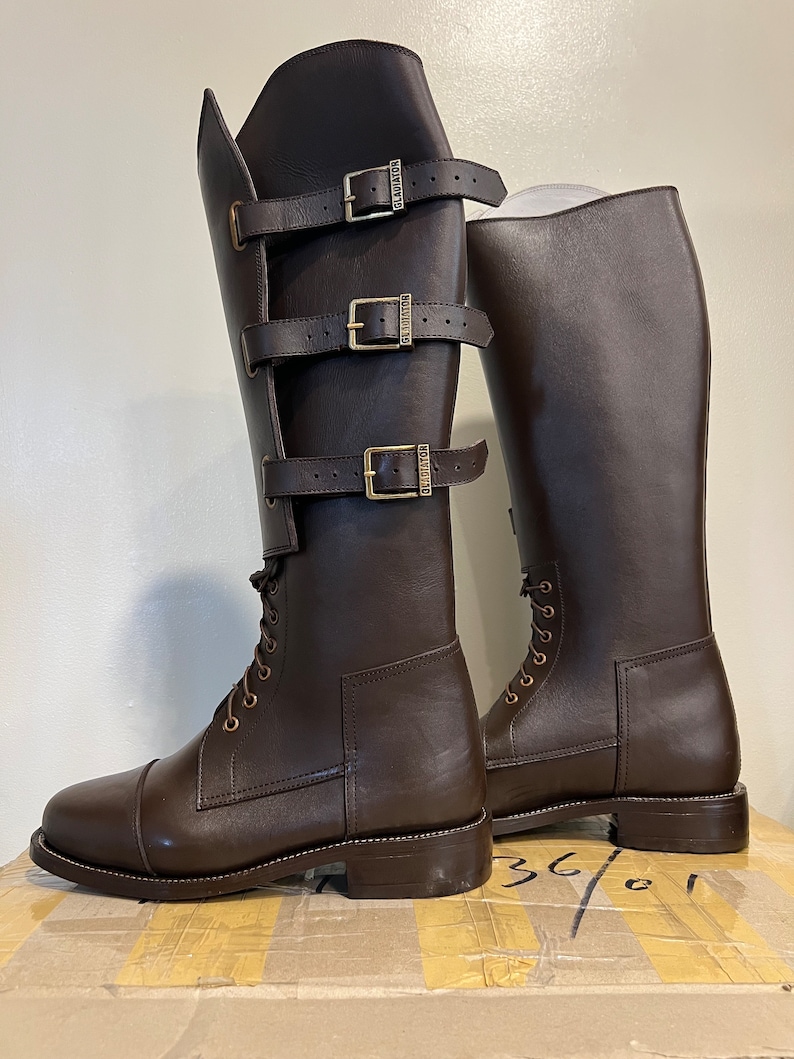 May include: A pair of dark brown knee-high boots featuring a lace-up front and a side panel secured by three horizontal straps with brass-toned buckles. Each buckle is embossed with the word GLADIATOR. The boots have a rounded toe and a low heel.