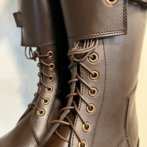 May include: A pair of dark brown leather boots featuring a tall shaft, a lace-up front with brass-toned metal eyelets, and a wide strap across the upper section secured with a matching brass-toned buckle closure for a structured fit.