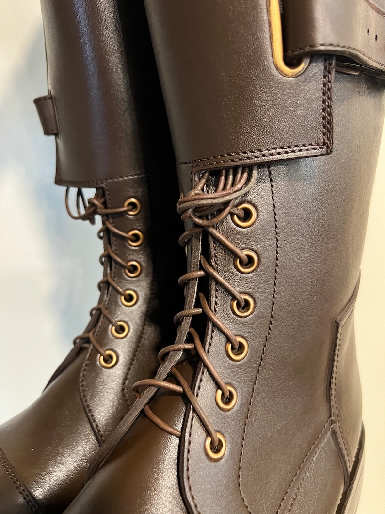 May include: A pair of dark brown leather boots featuring a tall shaft, a lace-up front with brass-toned metal eyelets, and a wide strap across the upper section secured with a matching brass-toned buckle closure for a structured fit.